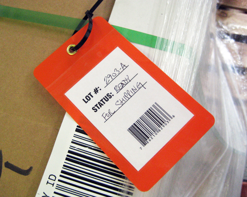 An image of an orange inventory tag with a barcode and hand written information shown attached to a carboard box of inventory products.