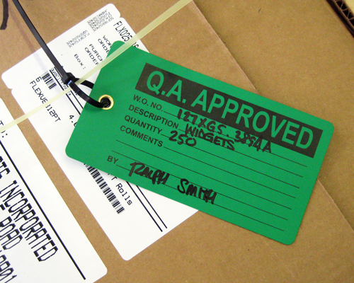 An image of a production status tag with black text on a green background reading, "Q.A. Approved". Tag is rectangular with rounded corners and a metal eyelet and is attached to a box.