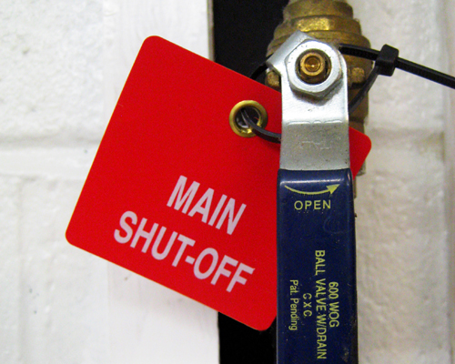 An image of a pre-printed plastic valve tag reading, "Main shut-off" in white on a red background. The tag is square with rounded corners and a metal eyelet and is attached to a valve.